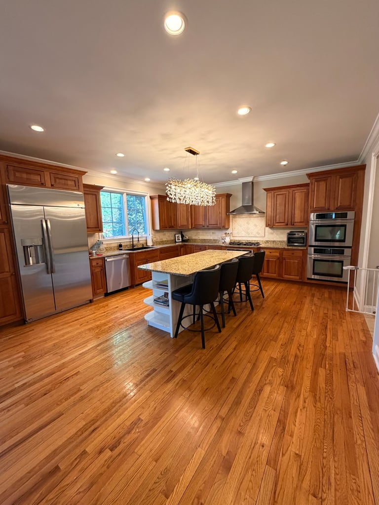 Spacious modern kitchen with wood cabinets, stainless steel appliances, marble island with seating, and wood flooring.