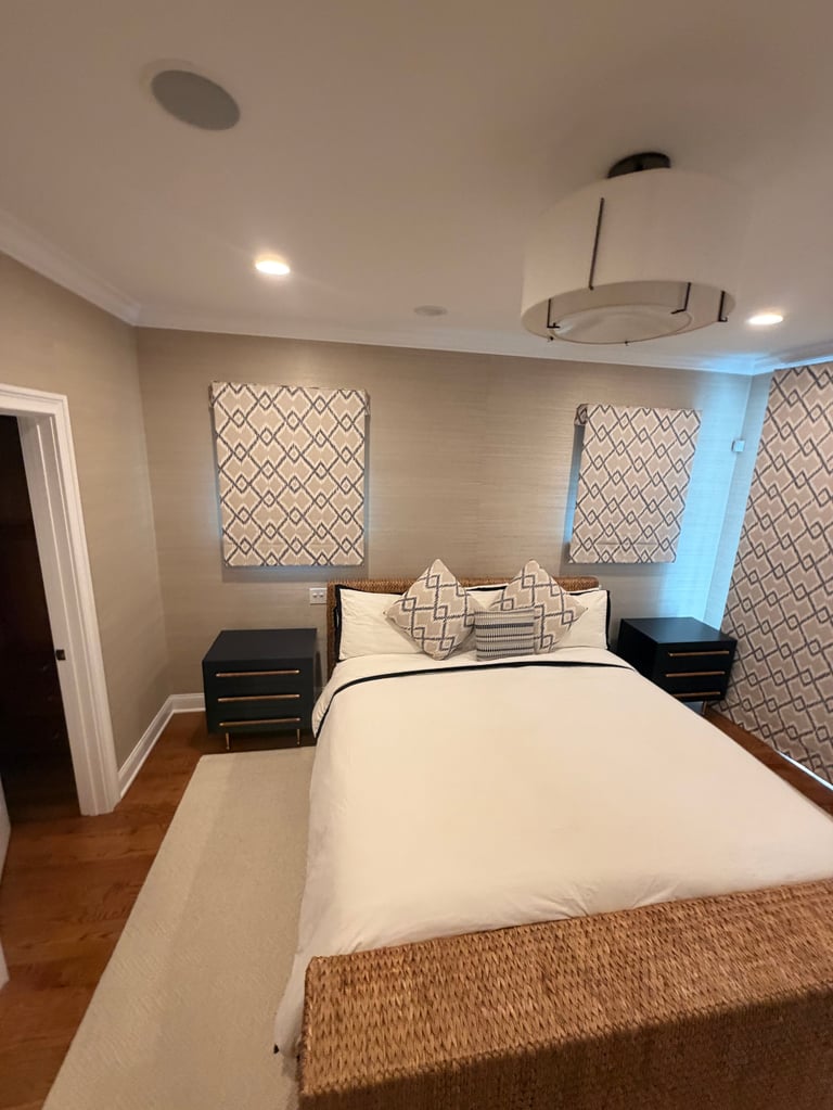 Modern bedroom with white bedding, geometric patterned window shades, black nightstands, and warm recessed lighting from ceiling fixtures.