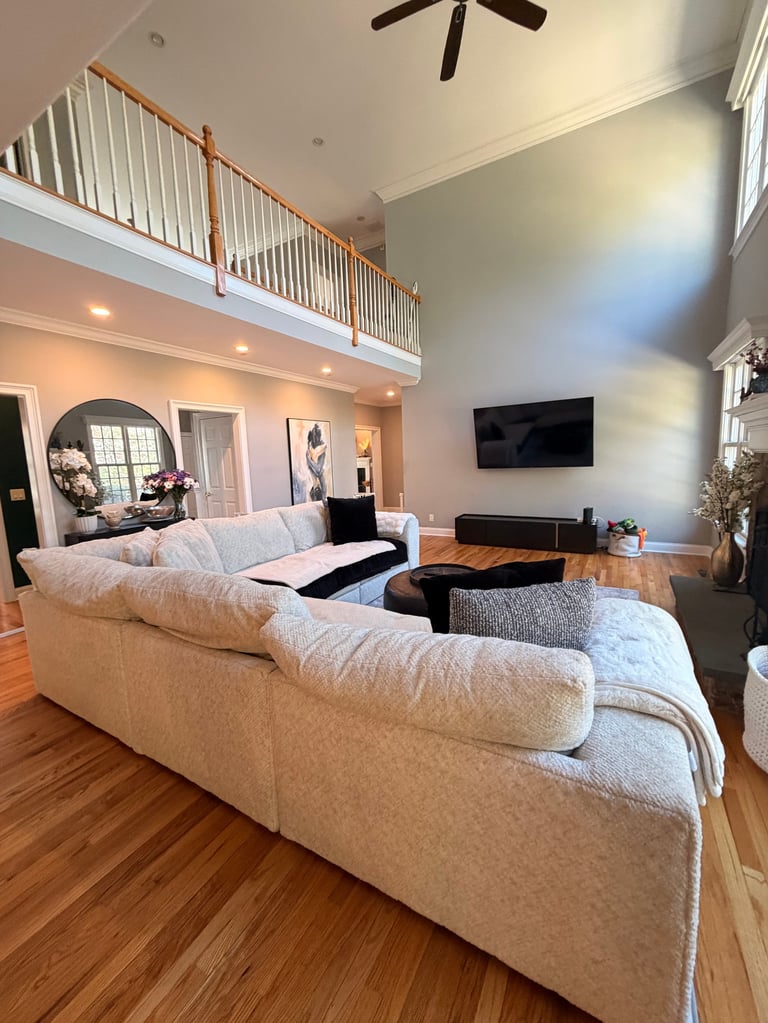 Modern living room with two beige sofas facing a wall-mounted TV, wooden floors, and a mezzanine loft area above with metal railings.