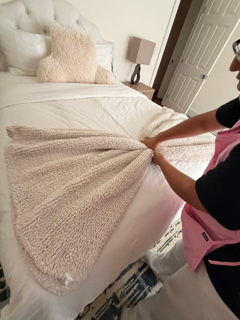 Person making bed with cream and beige textured blankets and pillows in bedroom