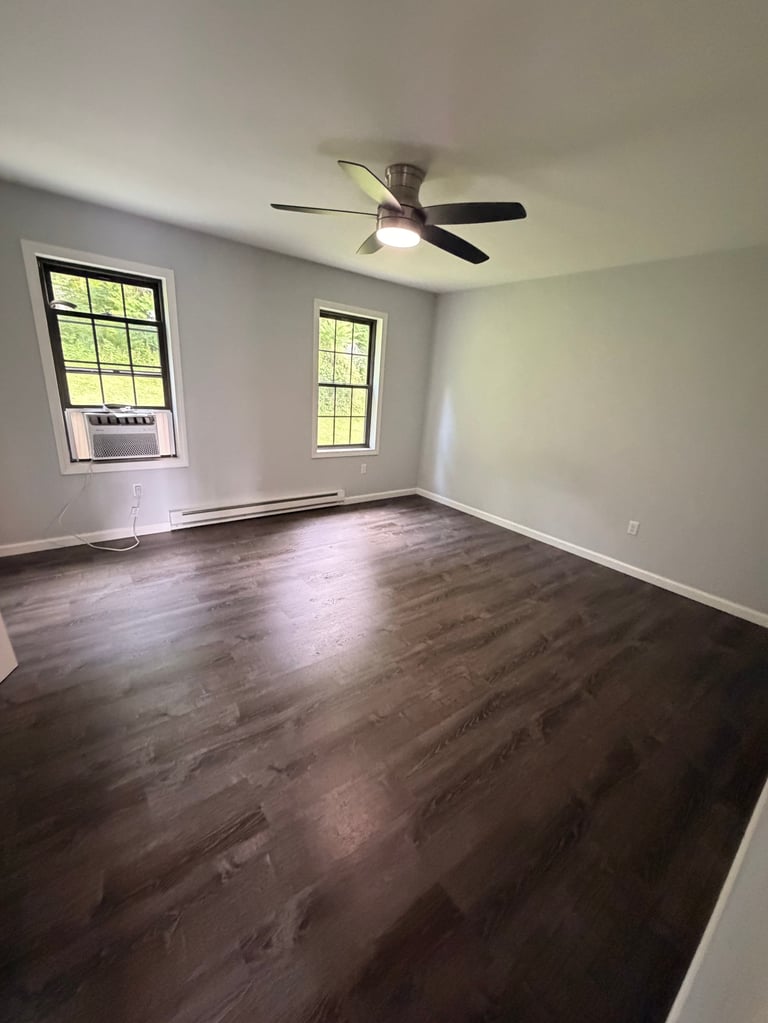 Empty room with dark wood flooring, white walls, two windows, and a ceiling fan with light fixture.