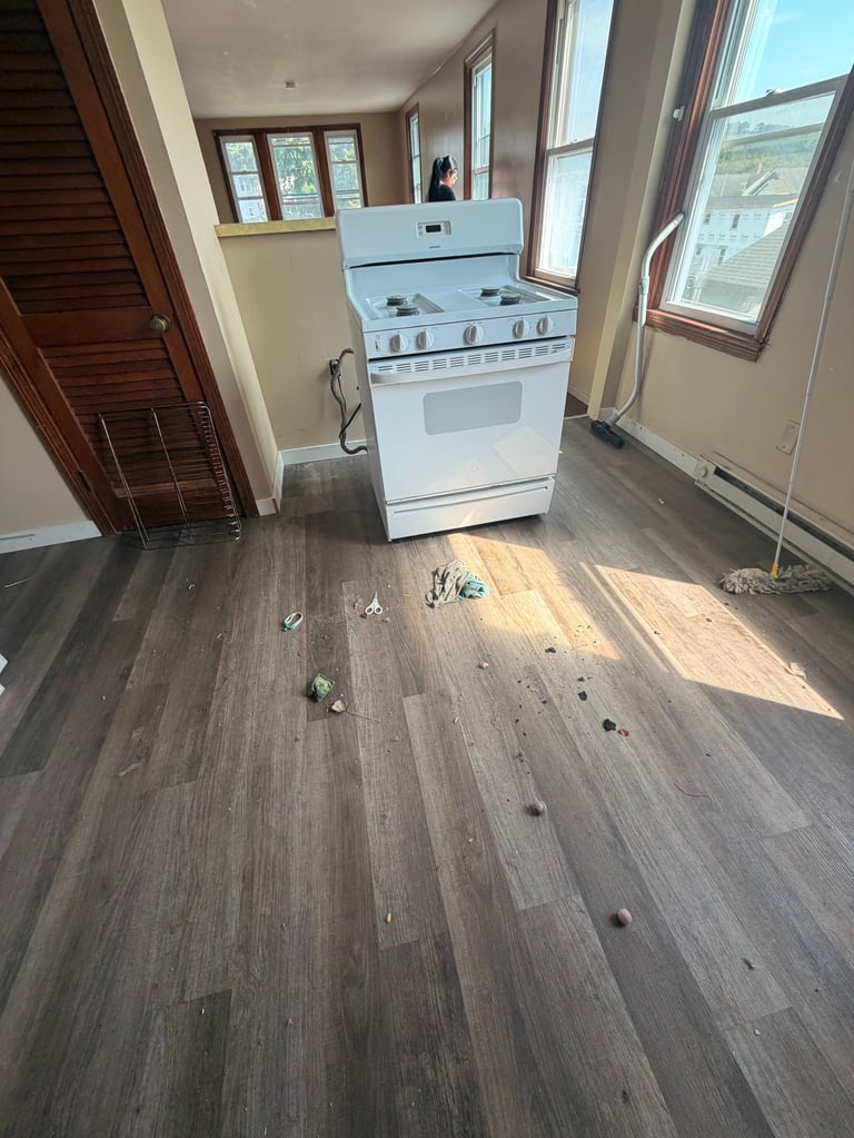 A white electric stove in a kitchen with gray laminate flooring under renovation, with windows and wooden trim visible.