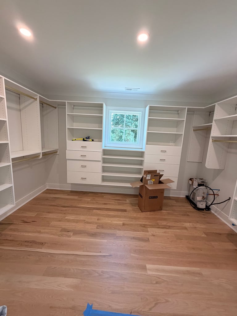 Spacious walk-in closet with white built-in cabinetry, wooden floor, window, and cardboard boxes on the floor