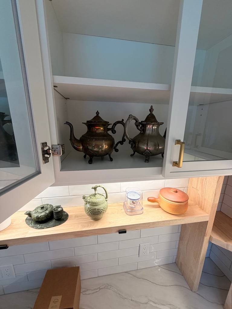 Kitchen nook with open shelving displaying ornate teapots above a wooden counter with small plants and kitchenware