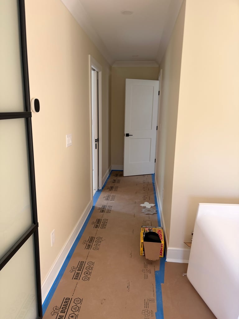 Long hallway with cream walls, white doors, cardboard subfloor covered with blue tape, and yellow measuring tool on the floor