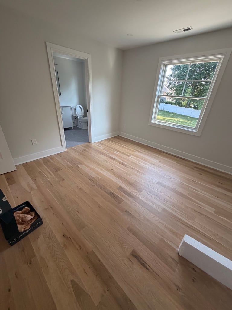 Empty room with light wood flooring, white walls, open doorway to bathroom, and large window with outdoor view