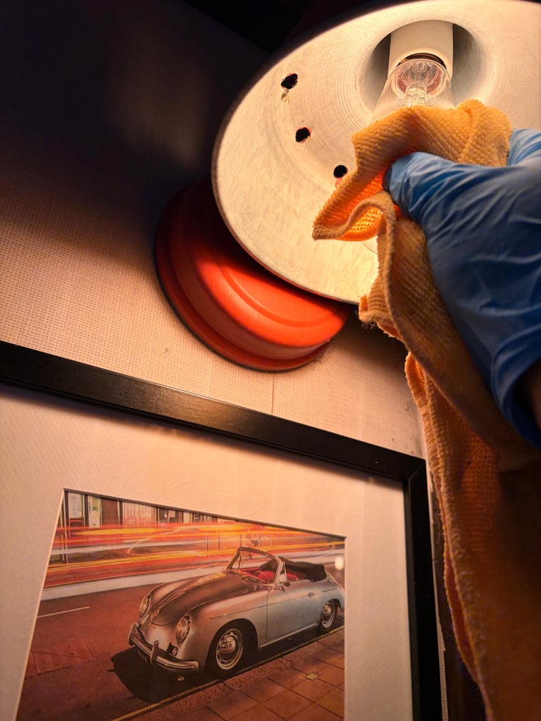 Hand holding orange cloth cleaning a round wall art piece with red accent, with vintage car photo visible below
