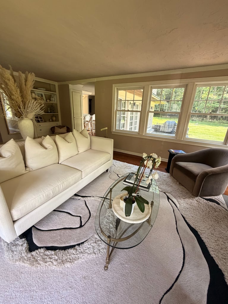Modern living room with cream sofa, glass coffee table with plant, brown armchair, and large windows overlooking green lawn
