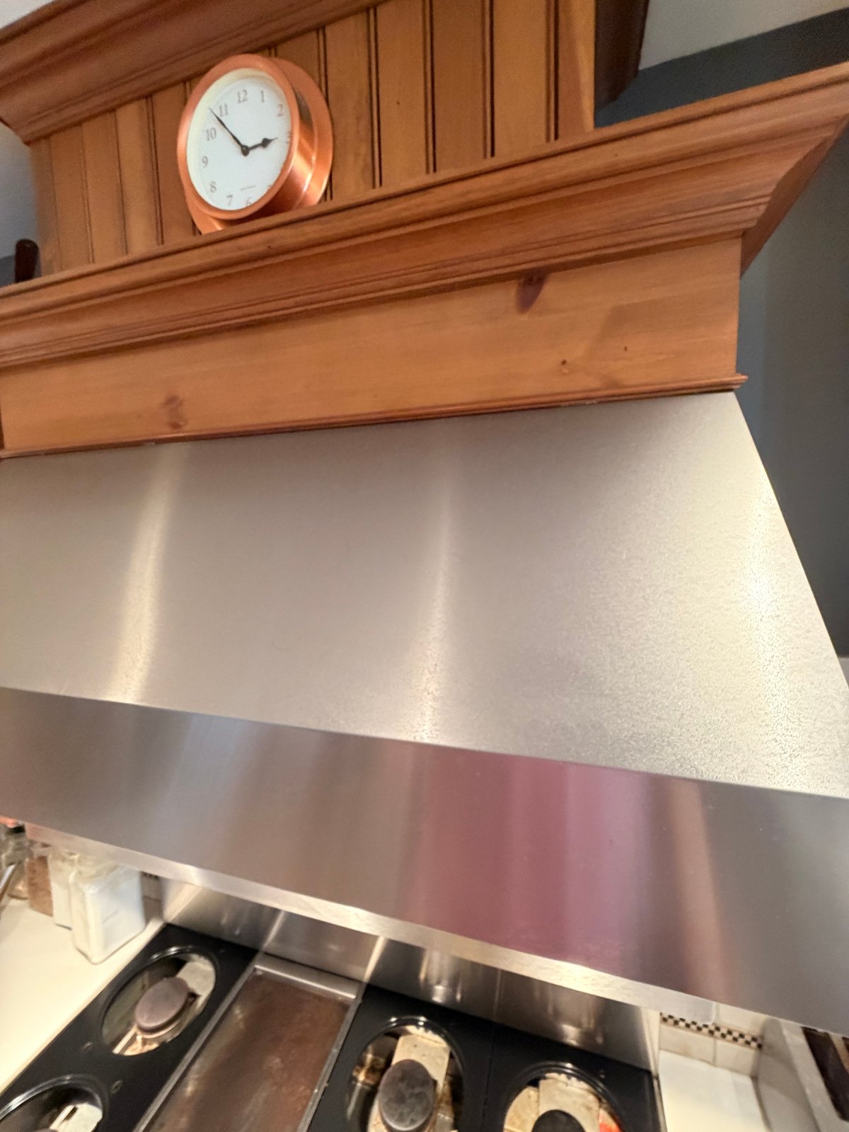 Modern kitchen range hood with stainless steel finish beneath wooden cabinetry and clock