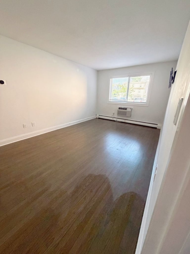 Empty apartment bedroom with hardwood flooring, white walls, large window, and wall-mounted air conditioning unit
