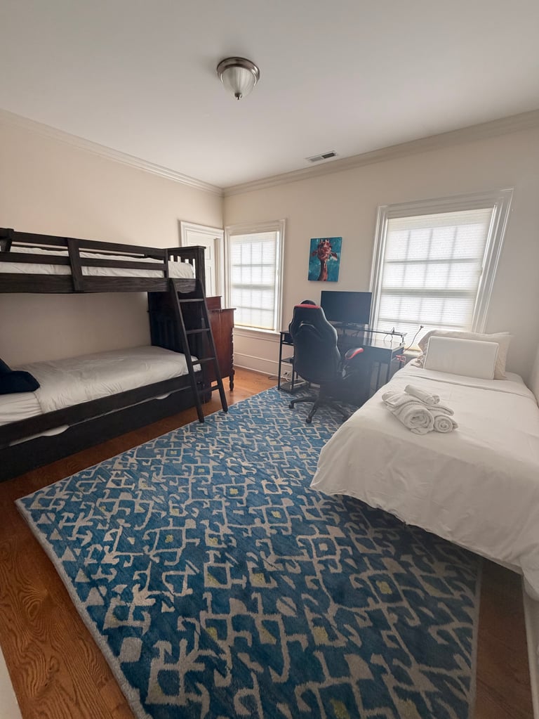 Modern dorm room with black bunk bed, single bed with rolled towels, desk, blue patterned rug, and two large windows.