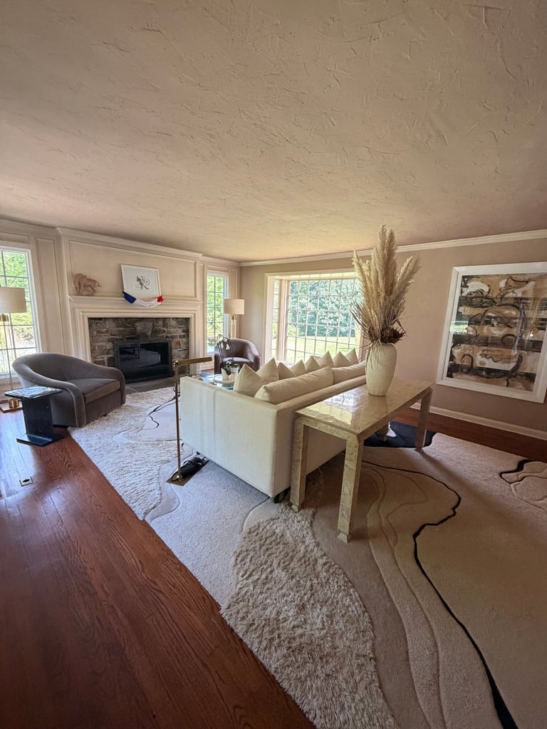 Spacious living room with cream sofa, dark armchair, fireplace, hardwood floors, area rugs, large windows, and decorative artwork.