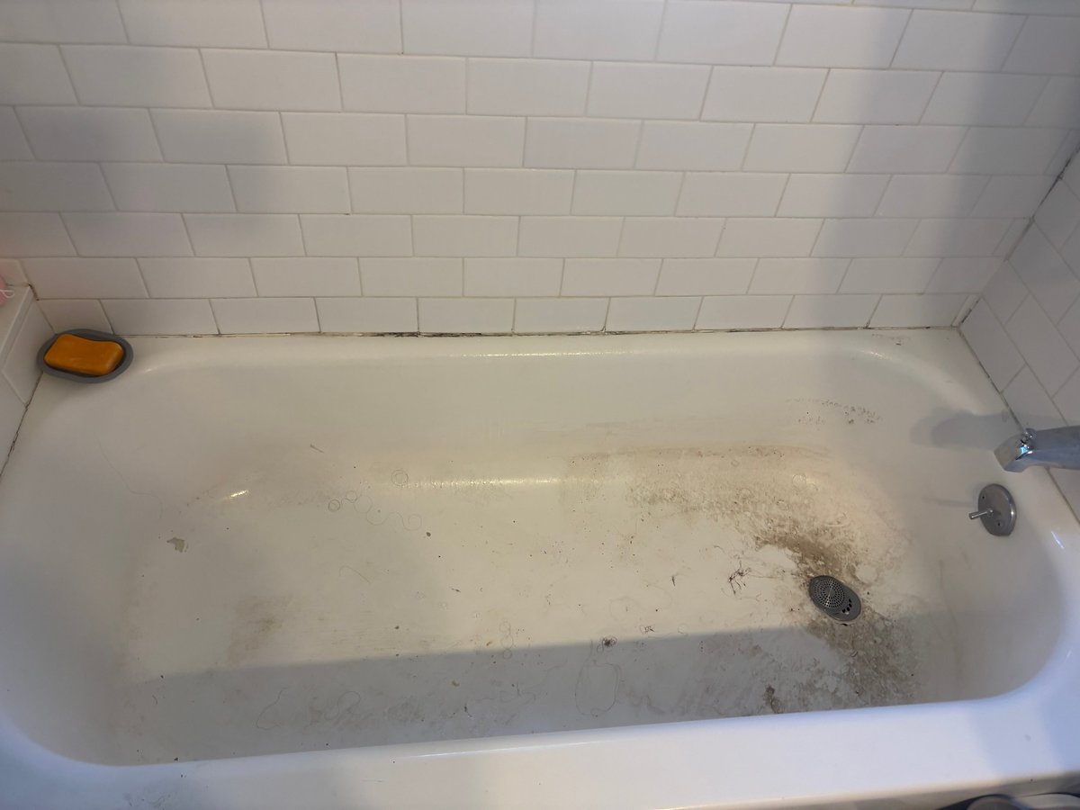 Dirty bathtub with stains and debris, white tile walls, and an orange soap dish on the edge