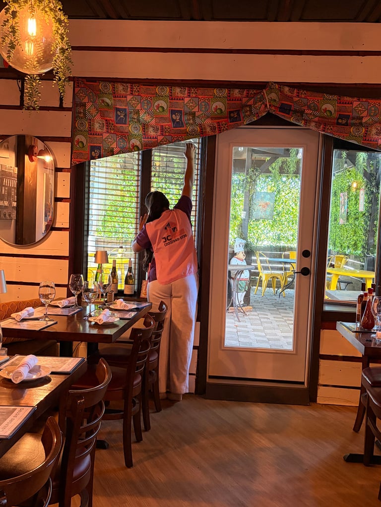 Woman in orange shirt standing in warm-lit restaurant dining room with tables, chairs, and large glass doors opening to an outdoor patio