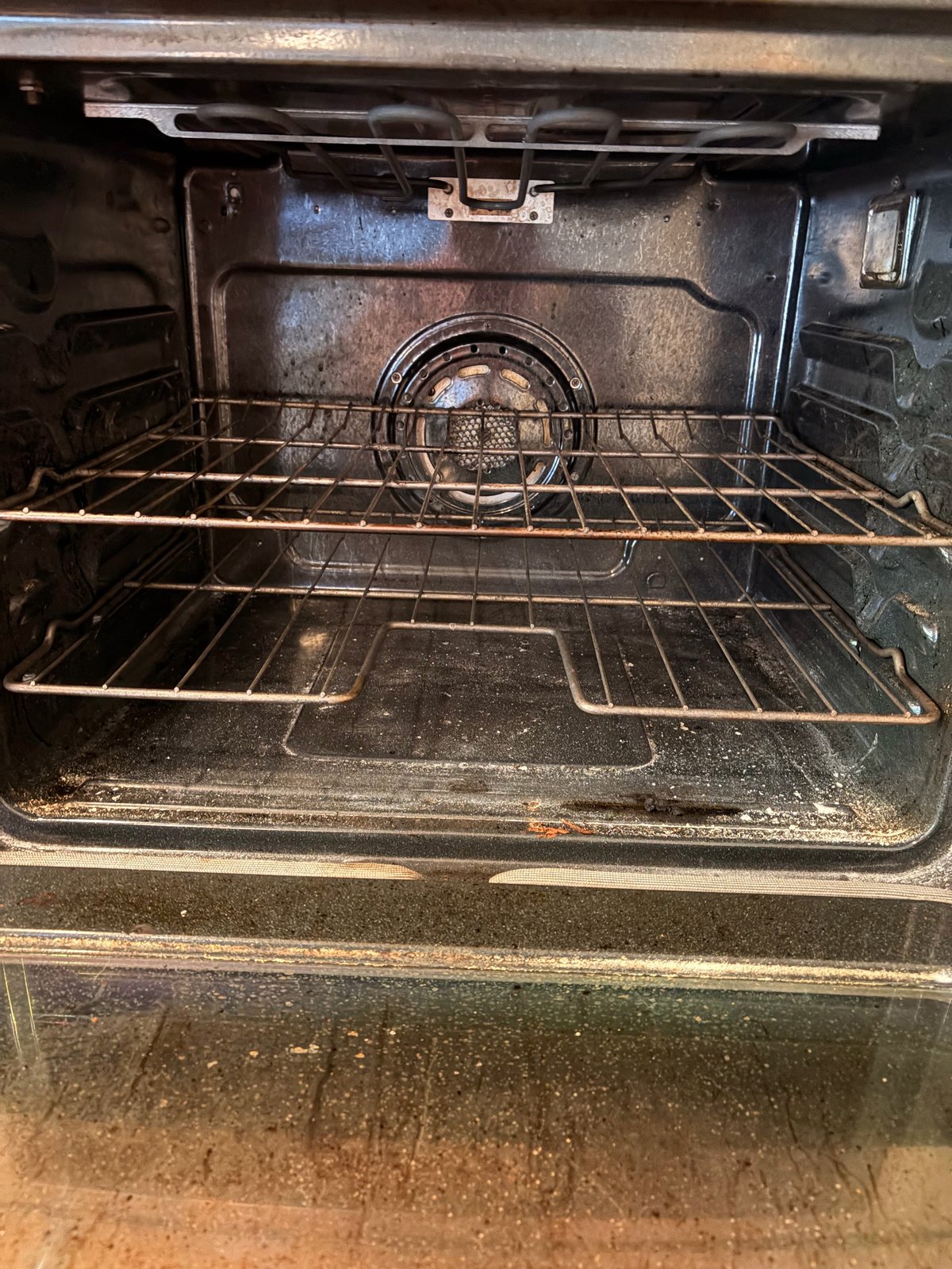 Interior view of a dirty oven with wire racks and a fan unit, showing buildup and grease on the sides and bottom surface