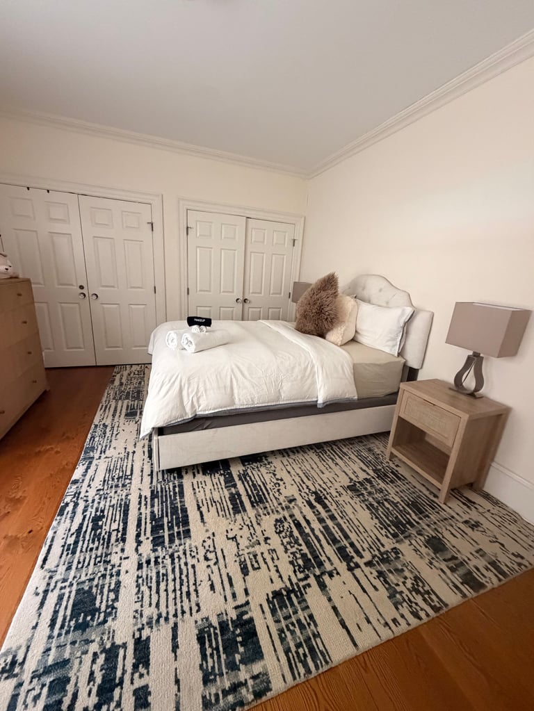 Bright, minimalist bedroom with a bed featuring white bedding and brown pillows, wooden nightstand, cream-colored walls, hardwood floors, and a geometric patterned area rug.
