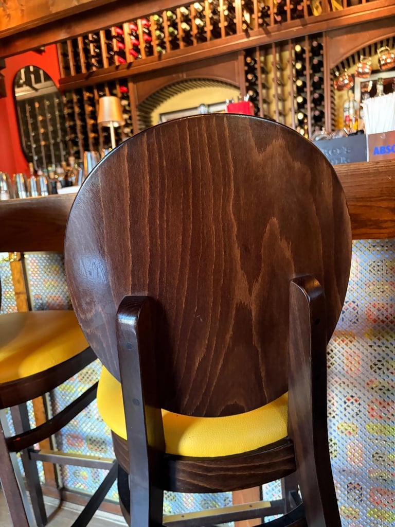 Wooden bar stool with dark brown back and yellow seat, viewed from behind in a restaurant or bar setting with wine bottles on shelves