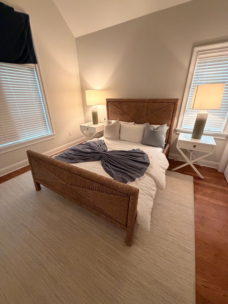 Bright bedroom with rattan bed frame, white bedding, dark throw blanket, two nightstands with lamps, and windows with shutters