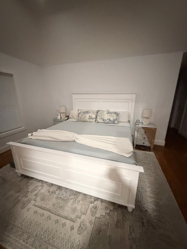 Bright bedroom with white furniture including a bed frame with patterned pillows, nightstands, and a bench at the foot of the bed on a geometric rug.