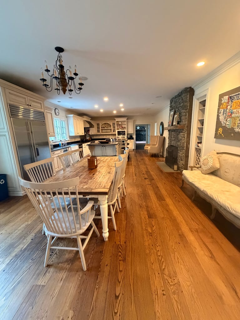 Open-concept home interior with wooden dining table, white chairs, hardwood floors, modern kitchen, and chandelier overhead.