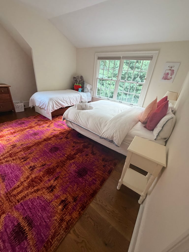 Bedroom with two white beds, purple-red patterned rug, glass doors opening to green trees, white walls, and wooden nightstand