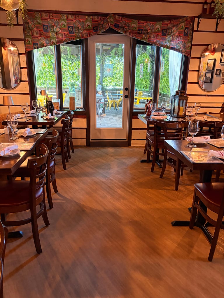 Elegant restaurant dining room with wooden tables, chairs, and hardwood flooring. Large glass doors open to a sunlit patio area visible in the background.