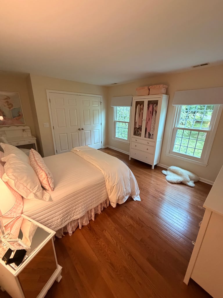 Bright, spacious bedroom with hardwood floors, white bed with pink pillows, cream walls, white closet doors, windows with natural light, and an armoire.